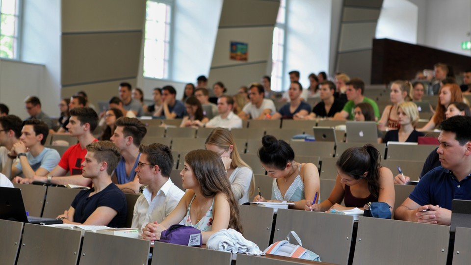 Studierende sitzen in einem Hörsaal. Die Studierenden tragen kurzärmlige Oberteile und vor ihnen liegen Laptops, Tablets oder Notizblöcke.