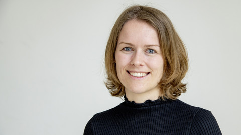 Laura Arnemann. She is wearing a black sweater and is standing in front of a greyish wall.