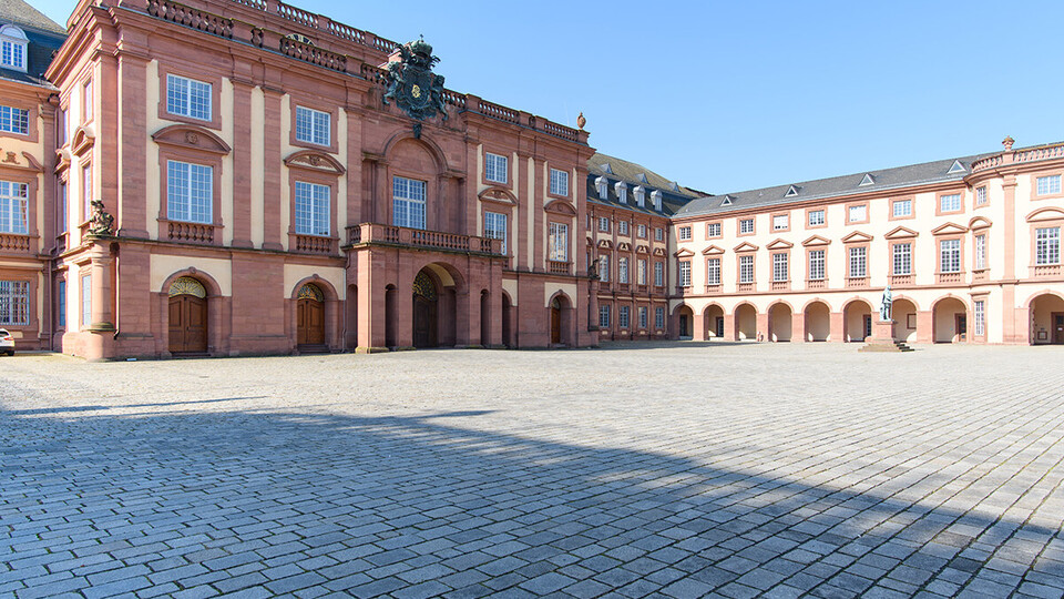 Die barocke Fassade des Mannheimer Schlosses im Ehrenhof, mit seinem markanten hellen Sandstein in Gelb und Rot und den symmetrisch angeordneten Fenstern und Bögen unter einem klaren blauen Himmel.