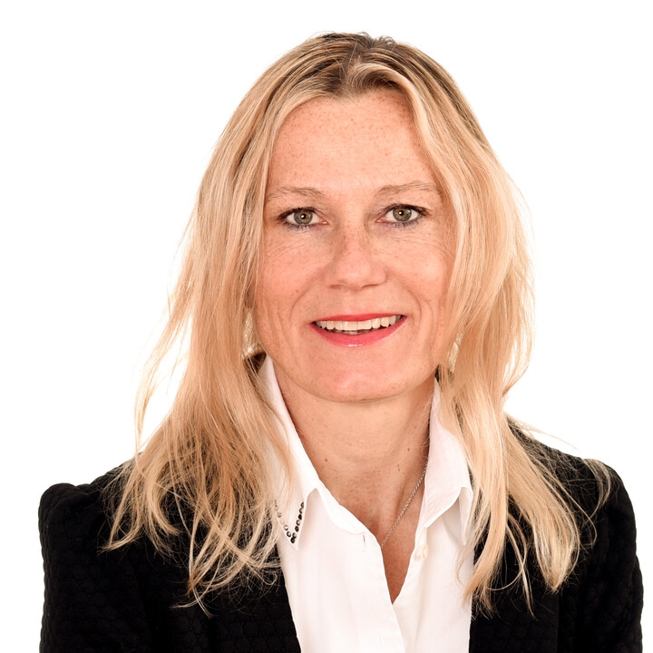 Portrait von Sabrina Scherbarth. Sie trägt eine weiße Bluse sowie einen schwarzen Blazer und lächelt.