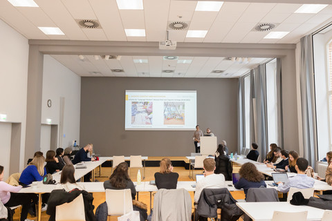 In einem Seminarraum sitzen einige Personen an Tischen. Zwei Personen stehen hinter einem Rednerpult und sprechen. An die Wand hinter dem Rednerpult wird eine Präsentation projiziert.