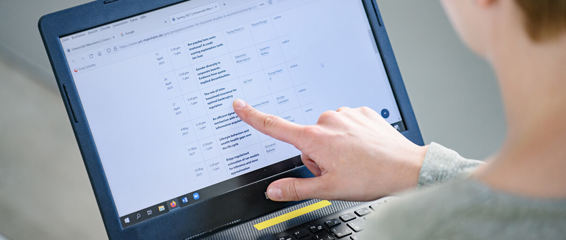 A doctoral student is holding a laptop and is pointing out a course on the screen where the schedule for different doctoral courses can be seen.