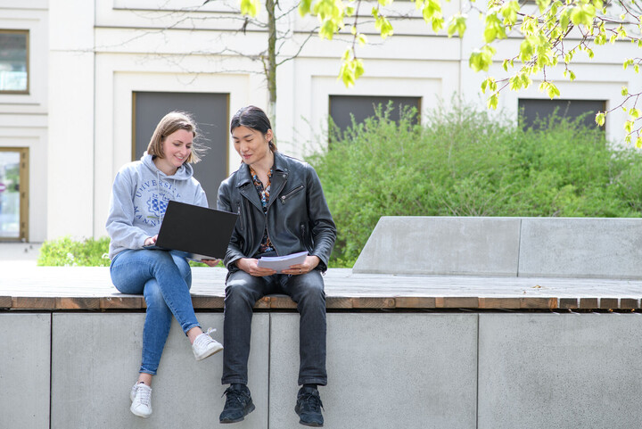 Zwei Studierende sitzen im Innenhof von B6. Sie haben einen Laptop auf dem Schoß und Karteikarten in der Hand.