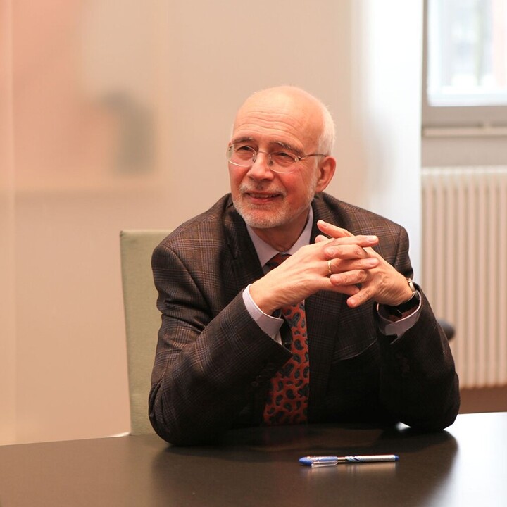Thomas Puhl trägt einen schwarzen Blazer und sitzt an einem Tisch. Ihm zugewandt sitzt eine Frau.