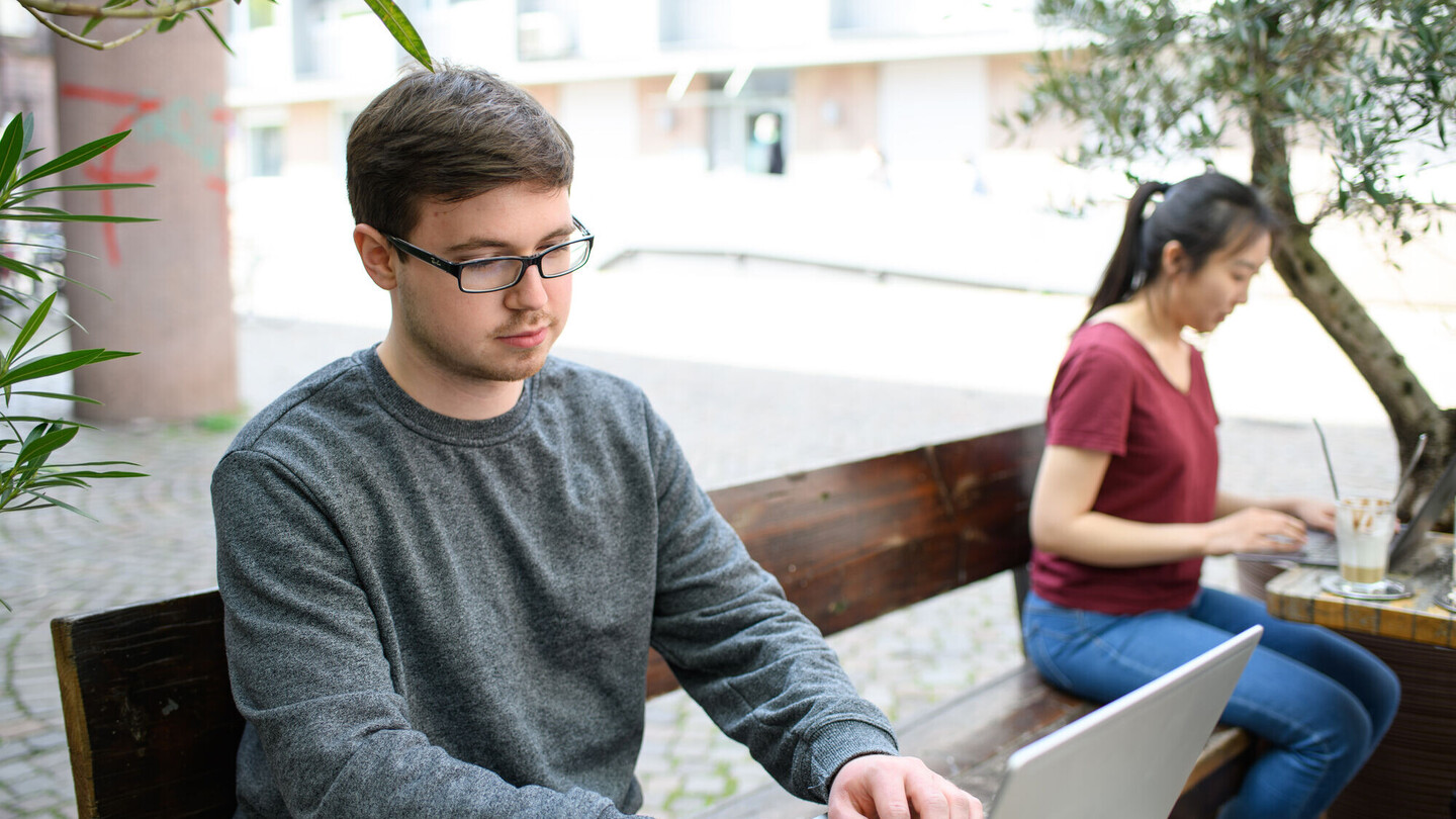 Zwei Studierende sitzen einzeln mit Laptop in einem Café auf einer Holzbank. Die vordere Person trägt einen grauen Pullover und hat kurze braune Haare, eine Brille und einen leichten Bartansatz. Die hintere Person hat lange schwarze Haare, die zu einem Pferdeschwanz zusammengebunden sind. Beide schauen konzentriert auf ihren Bildschirm.