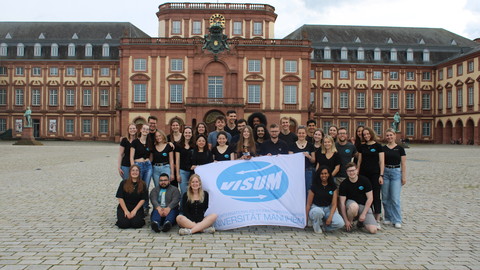 Eine Gruppe von Menschen steht auf dem Ehrenhof vor dem Schloss. Die halten ein weiße Fahne mit dem blauen Logo von "visum".