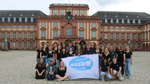 Eine Gruppe von Menschen steht auf dem Ehrenhof vor dem Schloss. Die halten ein weiße Fahne mit dem blauen Logo von "visum".