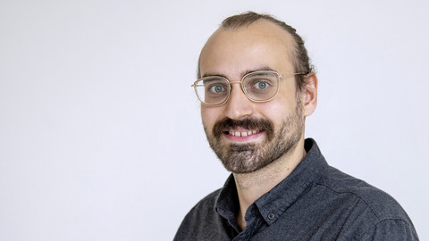 Alexander Kann. He is wearing a blackish shirt in front of a white background.