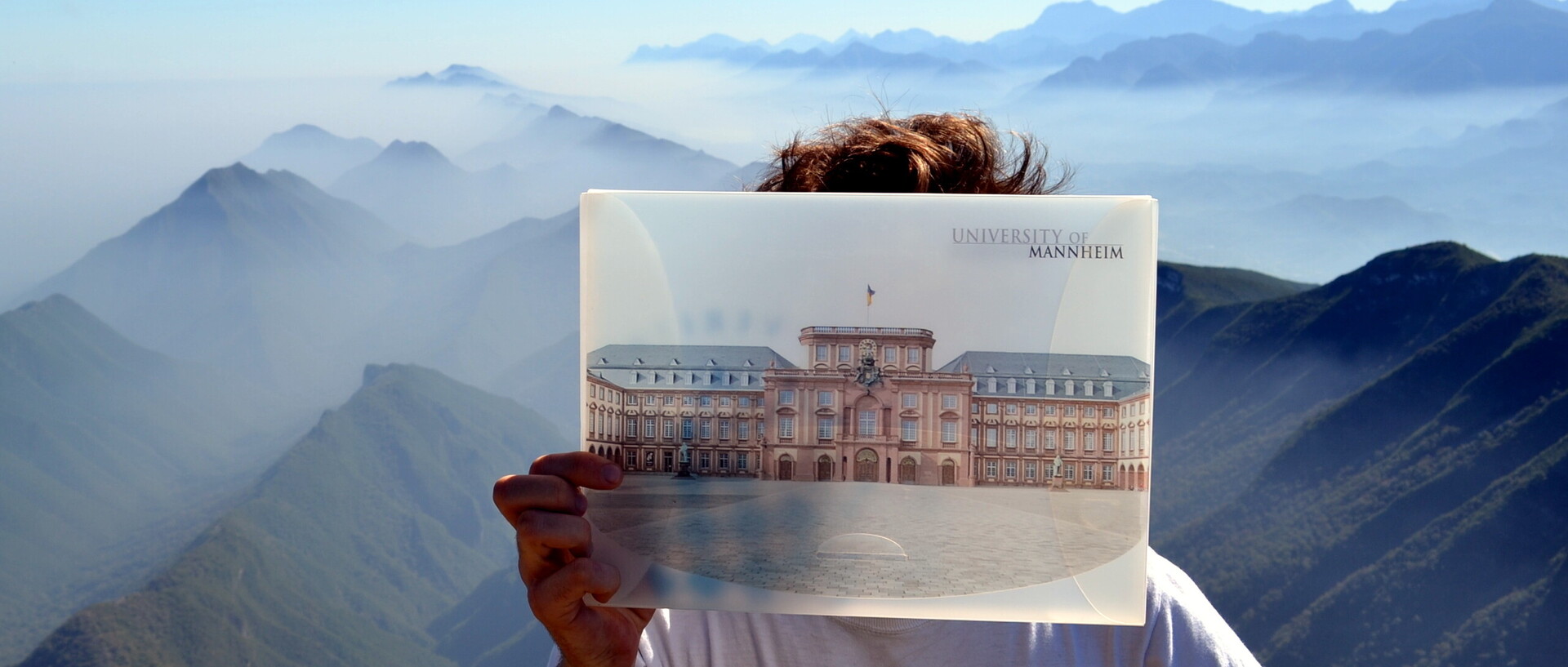Vor einer grünen, hügeligen Panoramalandschaft hält ein Student ein Bild von der Universität Mannheim vor sein Gesicht.