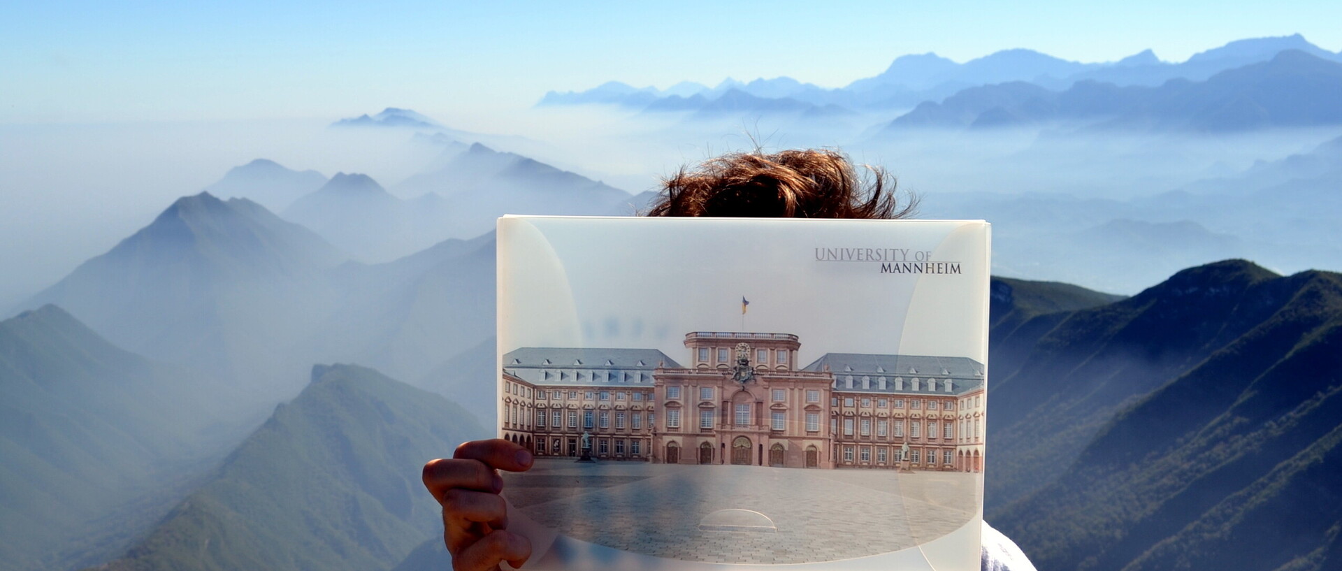 Vor einer grünen, hügeligen Panoramalandschaft hält ein Student ein Bild von der Universität Mannheim vor sein Gesicht.