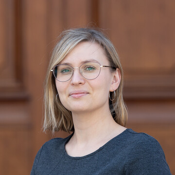 Maria Mühlbauer hat mittellange blonde Haare. Sier trägt eine runde Brille und ein dunkelgraues T-Shirt.