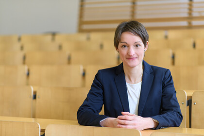 Portrait von Tabea Bucher-Koenen. Sie trägt eine weiße Bluse sowie einen dunkelblauen Blazer und sitzt mit gefaltetenen Händen in einem leeren Hörsaal.