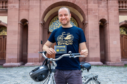 Mann hält einen Gutschein und steht mit Fahrrad im Ehrenhof vor dem Barockschloss.