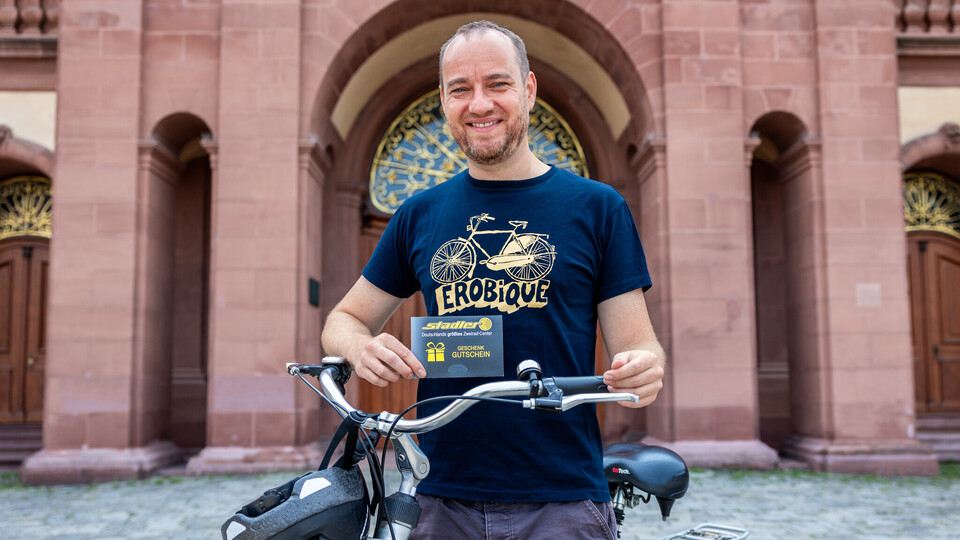 Mann hält einen Gutschein und steht mit Fahrrad im Ehrenhof vor dem Barockschloss.