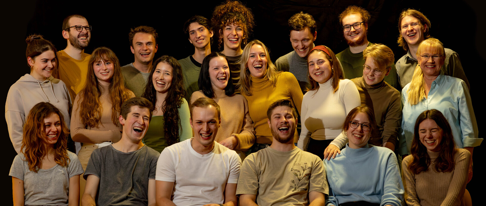 Eine Gruppe von mehr als 20 Menschen sitzt beziehungweise steht in drei Reihen hintereinander. Alle lachen ausgelassen.