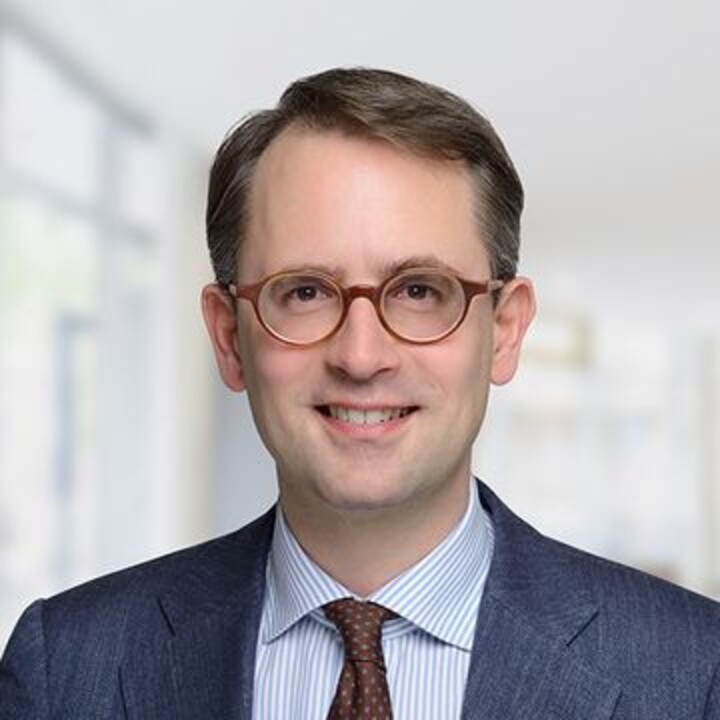Carsten Trenkler vor einem weißen Hintergrund. Er trägt eine Brille, Anzug, Krawatte und ein weißes Hemd.