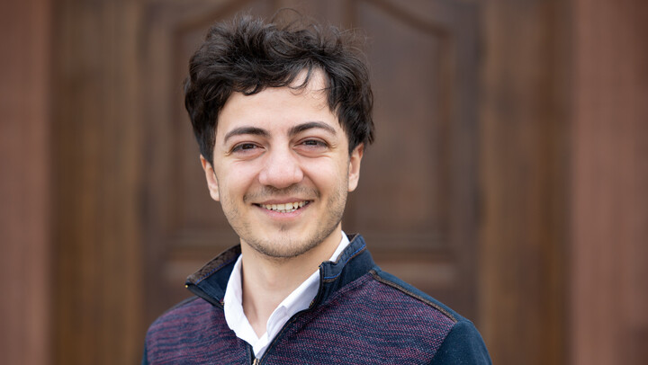 Argun Aman. He is wearing a purple jacket and is standing in front of brown door.