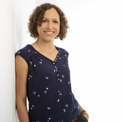 Isabel Thielmann. She is wearing a sleeveless blouse and is leaning against a white wall.