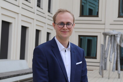 Thilo Dieing is standing in front of a university building. He has blond hair and is wearing glasses, a white shirt and a dark blue suit.