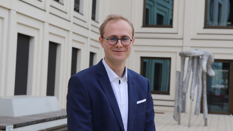 Thilo Dieing is standing in front of a university building. He has blond hair and is wearing glasses, a white shirt and a dark blue suit.