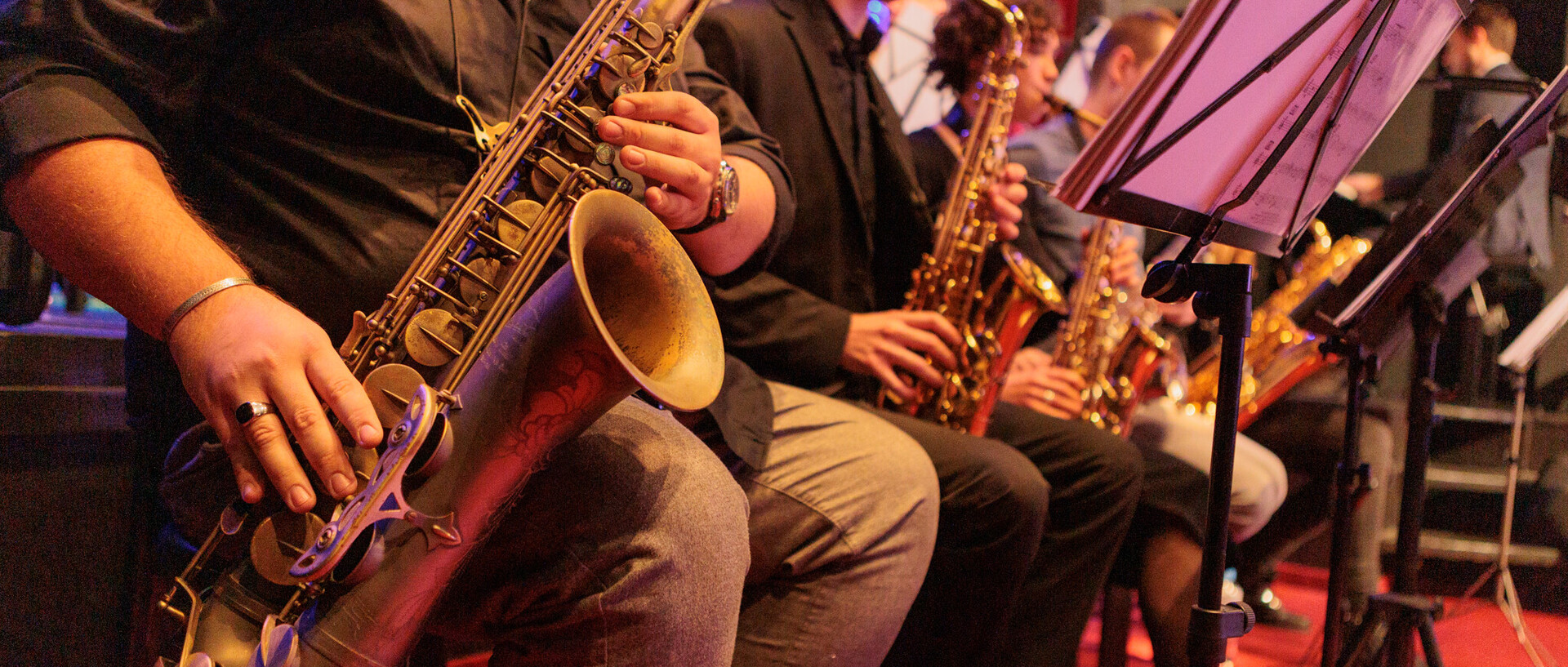 Eine Band sitzt auf Stühlen und spielt Blasinstrumente. Das Saxophon und die Hände des Saxophonisten sind scharf im Vordergrund, die anderen Instrumente unscharf im Hintergrund.