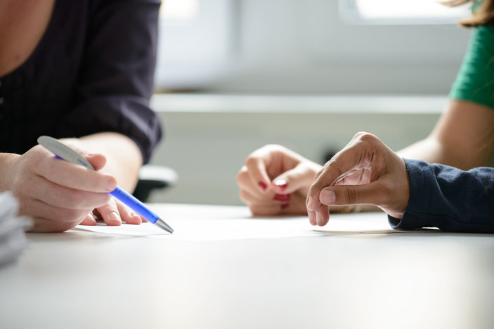 Zwei Studierende werden beraten, dabei sind die Hände und der Stift der Mitarbeiterin, der auf ein Papier zeigt, im Fokus.