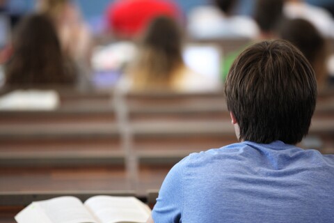 Rückenansicht eines Studierenden mit blauem T-Shirt und braunen Haaren. Er sitzt in einem Hörsaal, vor ihm ein aufgeklapptes Buch.