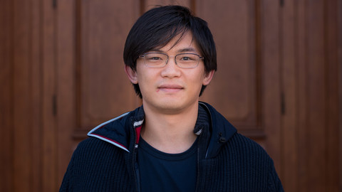 Taro Fujita. He is wearing a black jacket over a black shirt and is standing in front a brown door.