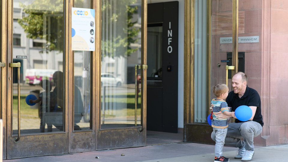 Eine erwachsene Person hockt vor dem Haupteingang zum Ostflügel der Universität Mannheim. Ein kleines Kind, das ihm gegenübersteht, schaut ihm direkt in die Augen und und hält dabei einen blauen Ballon. Beide scheinen in ein herzliches Gespräch vertieft zu sein.