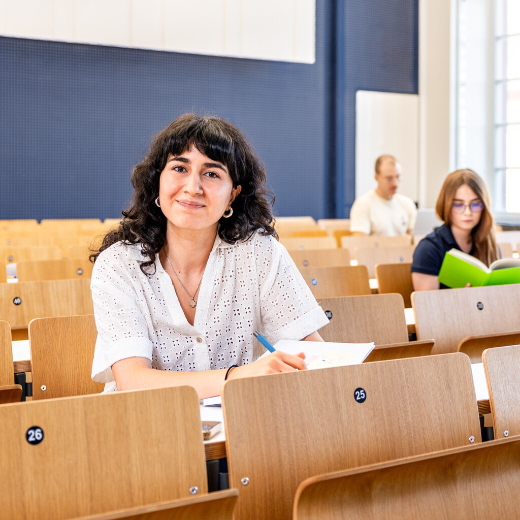 Eine Studentin sitzt in einer Bankreihe im Hörsaal. Sie schaut direkt in die Kamera und lächelt. Vor sich auf dem Tisch hat sie Unterlagen liegen. Im Hintergrund sitzen weitere Studierende an Laptops.