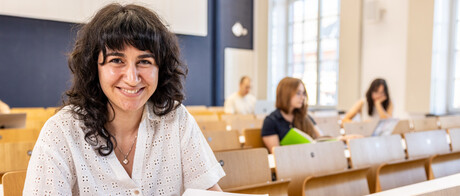 Eine Studentin sitzt in einer Bankreihe im Hörsaal. Sie schaut direkt in die Kamera und lächelt. Vor sich auf dem Tisch hat sie Unterlagen liegen. Im Hintergrund sitzen weitere Studierende an Laptops.
