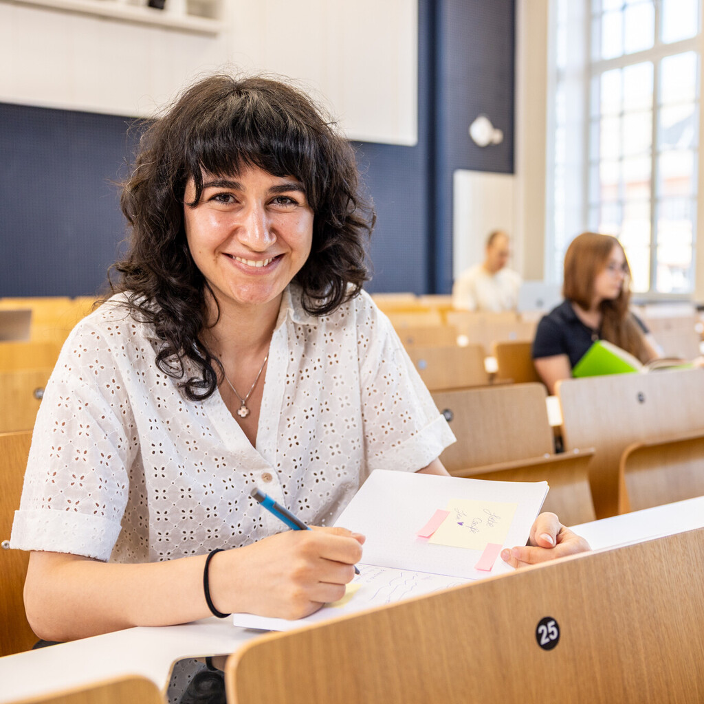Eine Studentin sitzt in einer Bankreihe im Hörsaal. Sie schaut direkt in die Kamera und lächelt. Vor sich auf dem Tisch hat sie Unterlagen liegen. Im Hintergrund sitzen weitere Studierende an Laptops.