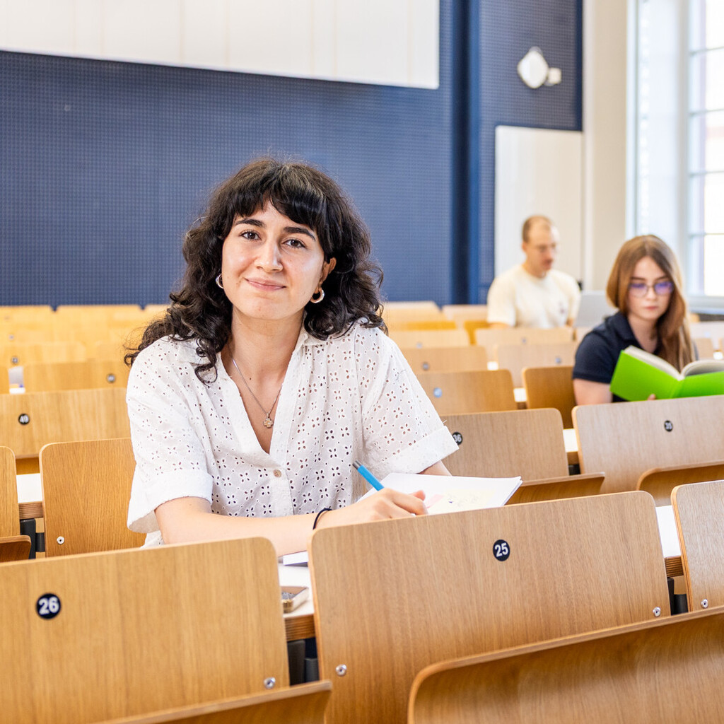 Eine Studentin sitzt in einer Bankreihe im Hörsaal. Sie schaut direkt in die Kamera und lächelt. Vor sich auf dem Tisch hat sie Unterlagen liegen. Im Hintergrund sitzen weitere Studierende an Laptops.