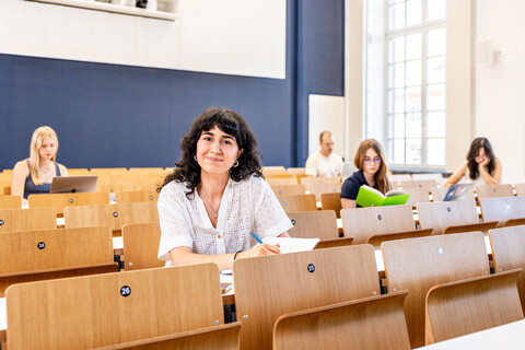 Eine Studentin sitzt in einer Bankreihe im Hörsaal. Sie schaut direkt in die Kamera und lächelt. Vor sich auf dem Tisch hat sie Unterlagen liegen. Im Hintergrund sitzen weitere Studierende an Laptops.