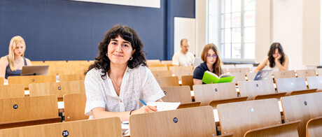 Eine Studentin sitzt in einer Bankreihe im Hörsaal. Sie schaut direkt in die Kamera und lächelt. Vor sich auf dem Tisch hat sie Unterlagen liegen. Im Hintergrund sitzen weitere Studierende an Laptops.