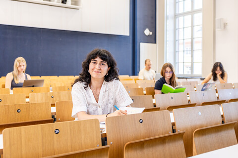 Eine Studentin sitzt in einer Bankreihe im Hörsaal. Sie schaut direkt in die Kamera und lächelt. Vor sich auf dem Tisch hat sie Unterlagen liegen. Im Hintergrund sitzen weitere Studierende an Laptops.