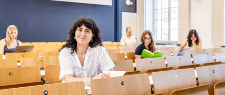 Eine Studentin sitzt in einer Bankreihe im Hörsaal. Sie schaut direkt in die Kamera und lächelt. Vor sich auf dem Tisch hat sie Unterlagen liegen. Im Hintergrund sitzen weitere Studierende an Laptops.