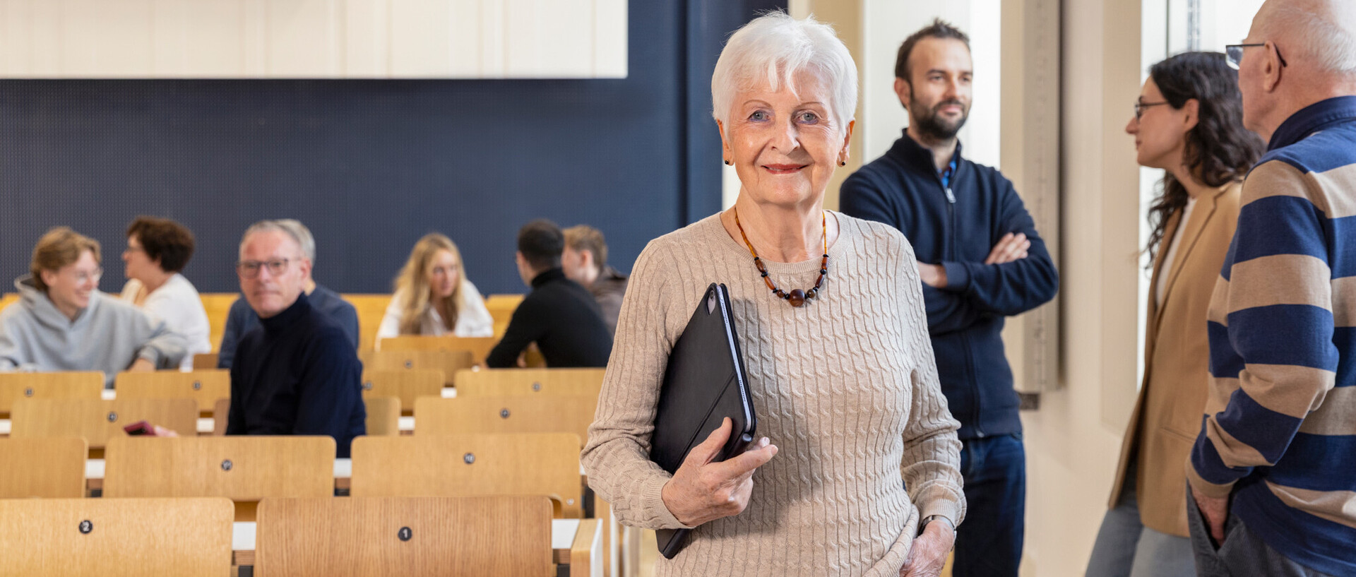 Eine Frau im höheren Lebensalter mit grauen Haaren schaut freundlich in die Kamera, im Hintergrund ein Hörsaal und sich unterhaltende Menschen verschiedener Altersstufen.