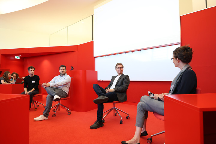 Vier Personen sitzen auf Drehstühlen in einem roten Seminarraum und führen eine Podiumsdiskussion. Sie tragen formelle Kleidung.