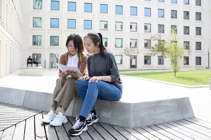 Two students are sitting and talking in the B6, 30-32 courtyard about doctoral studies.