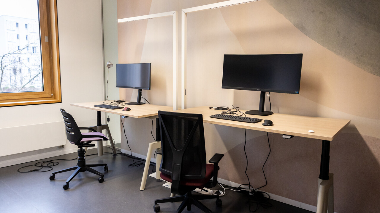 Das BIld zeigt zwei Arbeitsplätze in einem Doppelarbeitsraum des CAS. Auf den Tischen stehen geschwungene Bildschirme. Jeder Tisch verfügt über eine weiße rechtwinklige Leuchte.