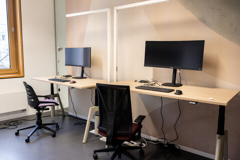 Das BIld zeigt zwei Arbeitsplätze in einem Doppelarbeitsraum des CAS. Auf den Tischen stehen geschwungene Bildschirme. Jeder Tisch verfügt über eine weiße rechtwinklige Leuchte.