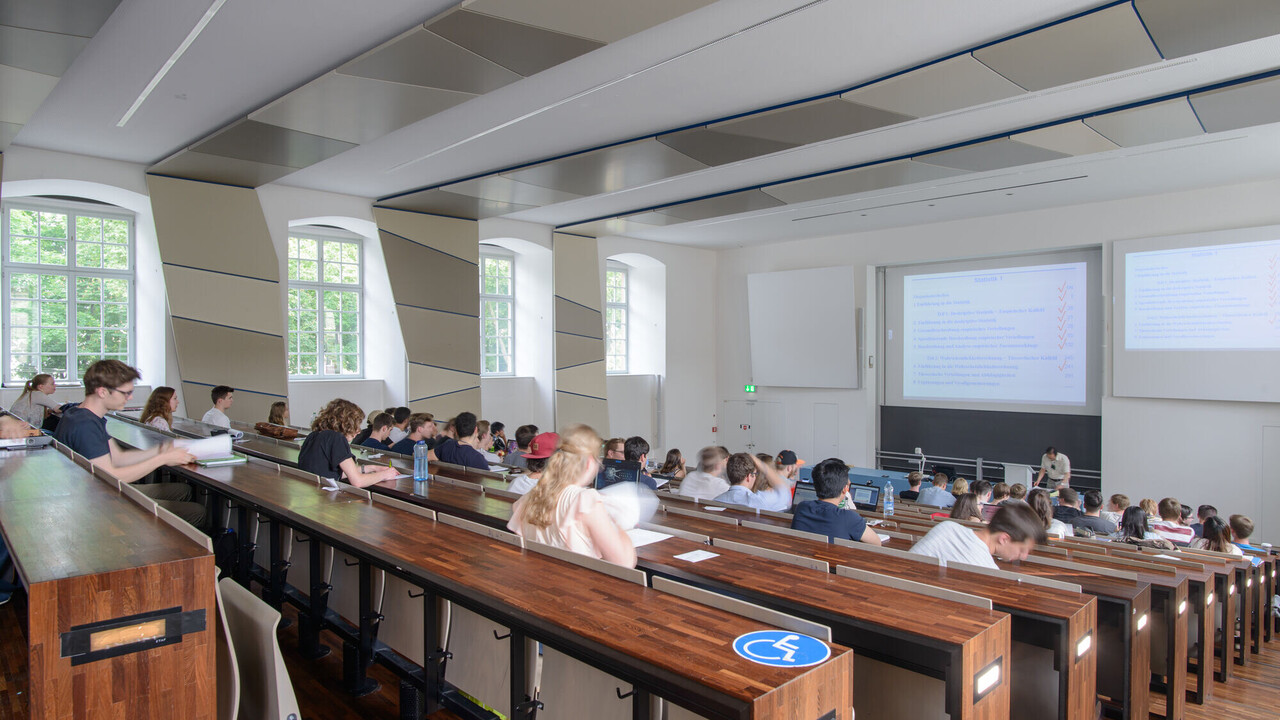 Studenten sitzen in einem Hörsaal und ein Professor hält eine Vorlesung.