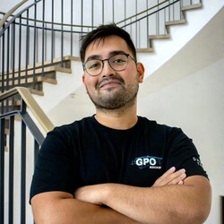 Christopher, mit kurzen schwarzen Haaren, Brille und schwarzem T-Shirt, lächelt mit verschränkten Armen freundlich in die Kamera.