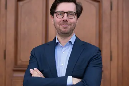 Die Person trägt eine Brille und ein dunkelblaues Sakko. Sie steht mit verschränkten Armen vor einer Holztür und lächelt.