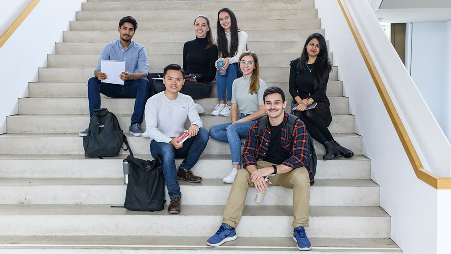 Eine Gruppe von sieben Studierenden sitzt auf einer hellen Treppe und lächelt in die Kamera. Sie haben Rucksäcke und Notizblöcke bei sich.