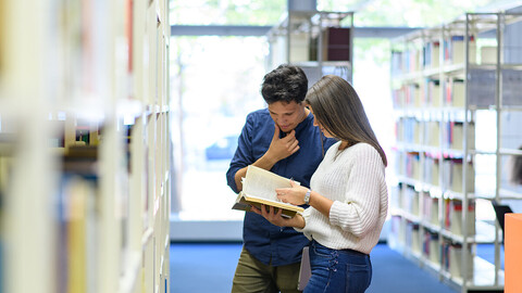 Zwei Studierende stehen vor dem Regal in der Bibliothek und schauen in ein Buch.