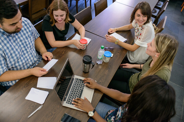 Vier Studentinnen und ein Student sitzen gemeinsam an einem Tisch. Sie unterhalten sich. Vor ihnen liegen Getränke, Karteikarten, Notizblöcke und ein Laptop.
