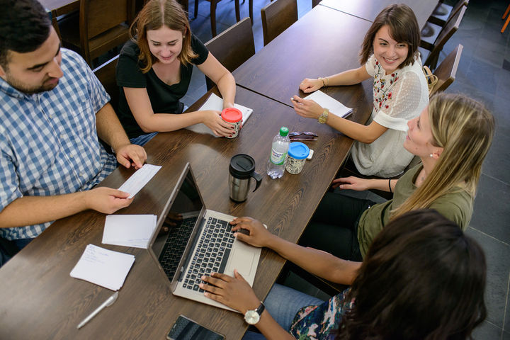 Vier Studentinnen und ein Student sitzen gemeinsam an einem Tisch. Sie unterhalten sich. Vor ihnen liegen Getränke, Karteikarten, Notizblöcke und ein Laptop.