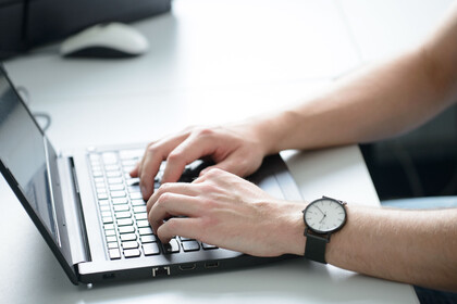 aufgeschlagener Laptop mit zwei Händen an der Tastatur, am rechten Handgelenk befindet sich eine Armbanduhr.